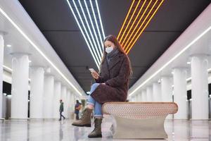 Eine Frau in einer medizinischen Gesichtsmaske wartet auf einen Zug und hält ein Smartphone in der Hand foto