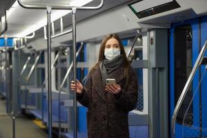 Eine Frau in einer Gesichtsmaske steht und benutzt ein Smartphone in einem modernen U-Bahn-Wagen