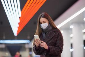 Eine Frau in einer medizinischen Gesichtsmaske wartet auf einen Zug und hält ein Smartphone in der Hand foto