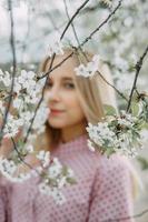 blond Mädchen auf ein Frühling gehen im das Garten mit Kirsche blüht. weiblich Porträt, Nahansicht. ein Mädchen im ein Rosa Polka Punkt Kleid. foto