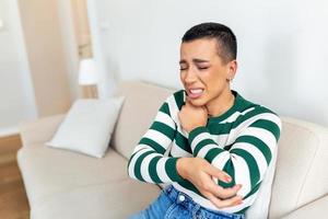 jung Frau ist Sitzung beim Zuhause auf ihr Sofa und berühren das ihr Ellbogen während Leiden von Ellbogen Schmerzen foto