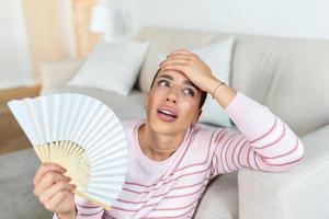 müde Frau Leiden von Hitzschlag oder heiß Sommer- eben ohne Klimaanlage, mit winken Fan, Sitzung im Leben Zimmer beim heim. Überhitzung foto