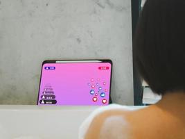 jung asiatisch Frau Aufpassen Leben Streaming über Tab Labor während Einweichen im das Badewanne. Aufpassen das Verkauf von Produkte online foto