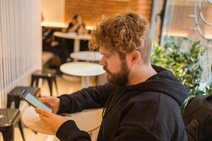 Porträt glücklich Kerl Herstellung Geld Transaktion über Anwendung auf Handy, Mobiltelefon Telefon mit 4g Internet Sitzung beim Cafe im Straße Abend, ernst tausendjährig Mann lesen Text Botschaft auf Smartphone foto