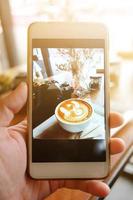 schön Hand halten Smartphone nehmen ein Foto und Bildschirm Schuss heiß Latté Kunst Kaffee mit Jahrgang Film Kamera und trocken Blume im Gras Vase mit natürlich sulight im Cafe