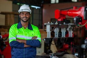 Porträt von ein afrikanisch amerikanisch Fabrik Arbeiter Arbeiten mit ein Roboter Arm im ein Fabrik. industriell Roboter Hintergrund zum automatisiert Produktion Technologie. suchen beim Kamera foto