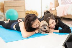 zwei schön asiatisch Mädchen tragen trainieren Kleider und lächelnd mit Hunde im Schlafzimmer. Frau auf Übung Matte mit Hanteln und Tablette foto