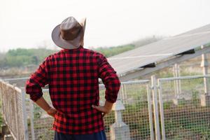 zurück Aussicht von asiatisch Mann Farmer trägt Hut, rot Plaid Shirt, stellen Hände auf Taillen, steht neben Solar- Panel draussen. Konzept, Farmer Eingerichtet Photovoltaik zum verwenden im Landwirtschaft zum Elektrizität. foto