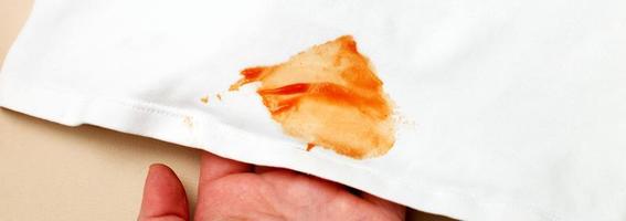 Frauen Hand halt Weiß Hemd Kleider mit Ketchup beflecken auf ein Beige Hintergrund. Banner foto