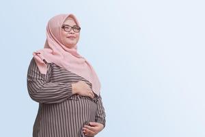 Porträt von schwanger asiatisch Frau mit hjiab halten Hände auf ihr Bauch oder Kind. zuerst Wochen von Schwangerschaft. isolieren Bild auf Blau Hintergrund. foto