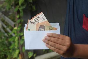 schließen oben von männlich Hand Einfügen fünf tausend Rupiah Geld Banknote in Umschlag. finanziell und Ersparnisse Konzept foto