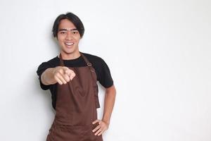 Porträt von attraktiv asiatisch Barista Mann im braun Schürze zeigen nach vorne zu das Kamera mit Finger. Werbung Konzept. isoliert Bild auf Weiß Hintergrund foto