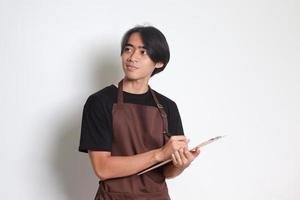 Porträt von attraktiv asiatisch Barista Mann im braun Schürze nehmen Befehl, Schreiben auf Speisekarte Buch Liste. isoliert Bild auf Weiß Hintergrund foto