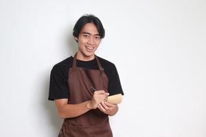 Porträt von attraktiv asiatisch Barista Mann im braun Schürze nehmen Befehl, Schreiben auf Speisekarte Buch Liste. isoliert Bild auf Weiß Hintergrund foto