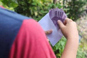 schließen oben von männlich Hand Einfügen zehn tausend Rupiah Geld Banknote in Umschlag. finanziell und Ersparnisse Konzept foto