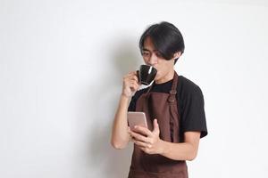 Porträt von attraktiv asiatisch Barista Mann im braun Schürze Trinken Kaffee während mit Handy, Mobiltelefon Telefon. isoliert Bild auf Weiß Hintergrund foto