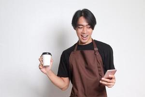 Porträt von attraktiv asiatisch Barista Mann im braun Schürze halten Einweg Papier Kaffee Tasse während mit Handy, Mobiltelefon Telefon. isoliert Bild auf Weiß Hintergrund foto