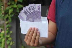 schließen oben von männlich Hand Einfügen zehn tausend Rupiah Geld Banknote in Umschlag. finanziell und Ersparnisse Konzept foto