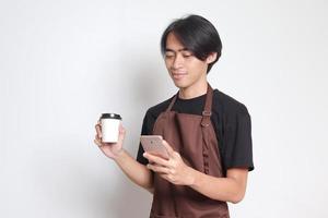 Porträt von attraktiv asiatisch Barista Mann im braun Schürze halten Einweg Papier Kaffee Tasse während mit Handy, Mobiltelefon Telefon. isoliert Bild auf Weiß Hintergrund foto