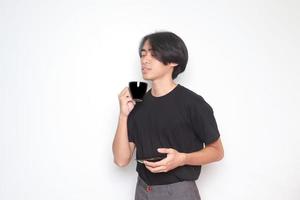 Porträt von gut aussehend asiatisch Mann im schwarz T-Shirt Trinken ein Tasse von Kaffee während halten das Untertasse. isoliert Bild auf Weiß Hintergrund foto