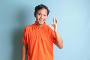 attraktiv asiatisch Mann im Orange Hemd zeigen in Ordnung Hand Geste und lächelnd suchen beim Kamera. isoliert Bild auf Blau Hintergrund foto