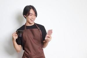 Porträt von attraktiv asiatisch Barista Mann im braun Schürze halten ein Tasse von Kaffee während mit Handy, Mobiltelefon Telefon und lächelnd beim Kamera. isoliert Bild auf Weiß Hintergrund foto
