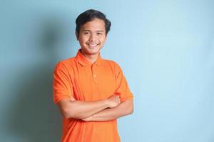 attraktiv asiatisch Mann im Orange Hemd behalten Waffen gekreuzt und suchen beim Kamera. isoliert Bild auf Blau Hintergrund foto