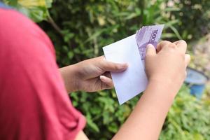 schließen oben von männlich Hand Einfügen zehn tausend Rupiah Geld Banknote in Umschlag. finanziell und Ersparnisse Konzept foto