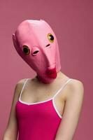 ein Frau tragen ein Rosa Fisch Kopf Maske auf Halloween steht und sieht aus beim das Kamera gegen ein Rosa Hintergrund. konzeptionelle Kunst Foto im ein verrückt Weg