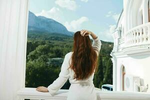 ein Frau im ein Weiß Kleid bewundert das Aussicht von das Berge von das Balkon halten ihr Haar foto