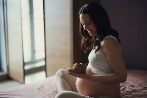 schwanger Frau entspannend beim heim. sie ist Sitzung auf Bett und halten Baby Schuhe. foto