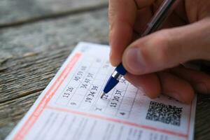 ein Mann ist Hand Kreuze aus das Zahlen im ein Lotterie Fahrkarte mit ein Stift auf ein hölzern Tisch. gewonnen. Glücksspiel Sucht, Sucht. foto
