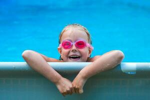 süß wenig Mädchen lächelnd im Brille im das Schwimmbad auf ein sonnig Tag. das Kind suchen Kamera . Sommer- Ferien foto