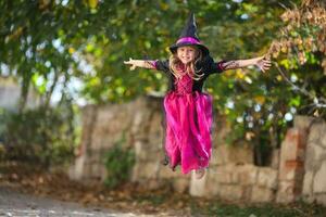 ein wenig Mädchen im ein Hexe Kostüm ist Springen auf das Straße. Halloween Urlaub. Kopieren Raum foto