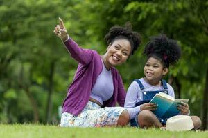 afrikanisch amerikanisch Lehrer ist Lehren ihr jung Schüler zu lesen während haben ein Sommer- draussen Klasse im das Öffentlichkeit Park zum Bildung und Glück Konzept foto