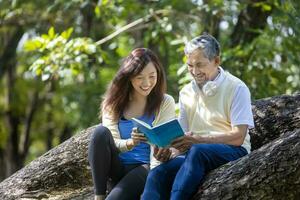 asiatisch Senior Vater und Tochter Sitzung und lesen ein Buch zusammen auf das Ast von reifen Baum während Sommer- zum physisch und mental Gesundheit und Langlebigkeit Verwendung. foto