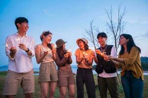 Gruppe von jung asiatisch Menschen sind genießen Camping , spielen Wunderkerze im natürlich Campingplatz beim Dämmerung foto