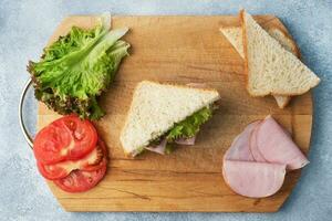 köstlich gesund Sandwich zum ein Snack Frühstück auf ein hölzern Schneiden Planke, oben Sicht. Toast Brot Tomate Schinken Blatt Salat. foto