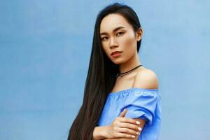 schön asiatisch Frau mit lange Haar im ein Blau Bluse Stehen in der Nähe von ein hell Blau Mauer foto