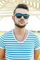 gut aussehend Mann im ein T-Shirt mit das Bart im Sonnenbrille Stehen auf das Strand. foto
