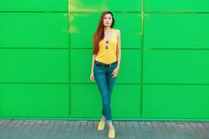 modisch schön Frau im stilvoll Gelb Kleider und Jeans Stehen in der Nähe von ein Grün Metall Mauer. foto