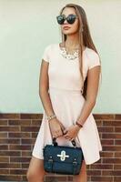 modisch schön Frau tragen Sonnenbrille im ein Rosa Kleid mit Handtasche foto