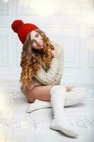 süß Frau mit lockig Haar im ein Jahrgang rot Hut, gestrickt Pullover und Socken Sitzung auf Weiß hölzern Fußboden im das Hintergrund von Weihnachten Beleuchtung foto