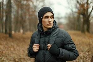 gut aussehend stilvoll jung Mann im Winter Jacke und Hut mit Rucksack ist Reisen im das Wald. foto