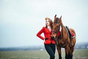 rothaariges Jockey-Mädchen in einer roten Strickjacke und schwarzen hohen Stiefeln mit einem Pferd foto