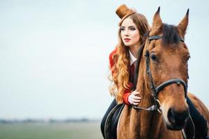 rothaariges Jockey-Mädchen in einer roten Strickjacke und schwarzen hohen Stiefeln mit einem Pferd