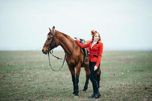 rothaariges Jockey-Mädchen in einer roten Strickjacke und schwarzen hohen Stiefeln mit einem Pferd foto