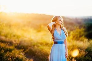 blonde Frau mit losen Haaren in einem hellblauen Kleid im Licht des Sonnenuntergangs foto