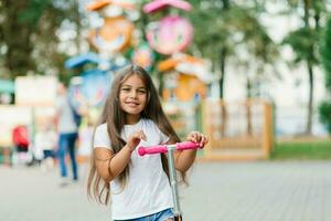 schön lächelnd Mädchen Fahrten ein Roller entlang das Pfad von ein Amüsement Park auf ein Sommer- Tag. saisonal Kinder- aktiv Sport. foto