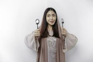ein asiatisch Muslim Frau ist Fasten und hungrig und halten Besteck während suchen beiseite Denken Über Was zu Essen foto